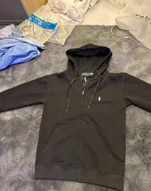Svart hoodie från Polo Ralph Lauren - Svart hoodie från Polo Ralph Lauren med klassisk logga på bröstet. Tröjan har huva med snörning, dragkedja framtill och två stora fickor. Perfekt för chill dagar och enkel att matcha med jeans eller joggers.