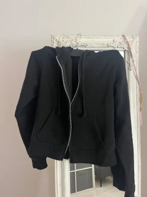 Svart hoodie med dragkedja - Svart hoodie med dragkedja och huva. Tröjan har två stora fickor framtill och snörning i huvan. Perfekt för chill dagar eller när du vill ha en enkel och stilren look. Materialet känns mjukt och bekvämt.