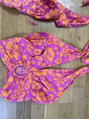 Blommig bikini från Bik Bok - Superfin topp från Bik Bok med tropiskt blommigt mönster i orange och rosa. Toppen har breda knytband och en snygg ringdetalj framtill på toppen. Materialet känns lätt och bekvämt, perfekt för strandhäng. Färgerna är riktigt somriga och sticker ut! (Hr två storlekar en o S&XS) 