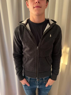 Svart hoodie från Polo Ralph Lauren - Svart hoodie från Polo Ralph Lauren med klassisk logga broderad på bröstet. Tröjan har huva, dragkedja framtill och två fickor. Insidan är ljusgrå och mjuk, perfekt för kyliga dagar. Snygg och enkel design som funkar till jeans och sneakers.