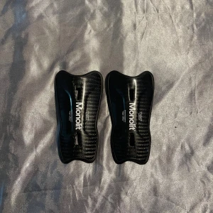 Monolit benskydd - Säljer ett par svarta benskydd från Monolit i kolfiber-look. De har en snygg, sportig form och är lätta men ändå stabila. Perfekta för dig som vill ha stilrena och funktionella benskydd till fotbollen. Sitter skönt mot benet och har en modern design.