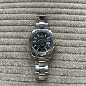  Sky-Dweller silver klocka -  Sky-Dweller med silverfärgad länk i metall och svart urtavla. Klockan har en rund boett, räfflad bezel och tydliga indexmarkeringar. Länken är robust och har -loggan på spännet. Perfekt för dig som gillar stilrena och lyxiga accessoarer.