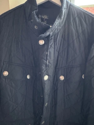 Jacka från Barbour M - Quiltad mörkblå jacka från Barbour i storlek M. Klassisk look med tryckknappar, ståkrage och två stora fickor framtill. Jackan har Barbour-logga på armen och är tillverkad i lätt polyester, perfekt för höst och vår.