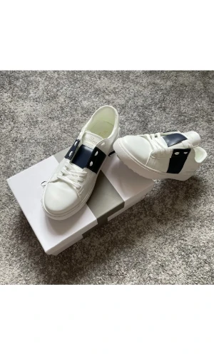 Valentino Sneakers - Snygga vita sneakers från Valentino Garavani med bred mörkblå rem och coola nitar på sidan. Skorna har rund tå, platt sula och klassisk snörning. Tillverkade i skinn för en lyxig känsla och stilren look. Perfekta för dig som gillar exklusiva märken och modern design.