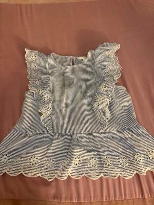 Randig blus med spets från H&M - Supersöt blus från H&M i storlek S med blåvita ränder och detaljerad vit spets längs volangärmar och nederkant. Blusen har en rund hals och är ärmlös, perfekt för en fräsch och somrig look. Materialet känns lätt och luftigt.