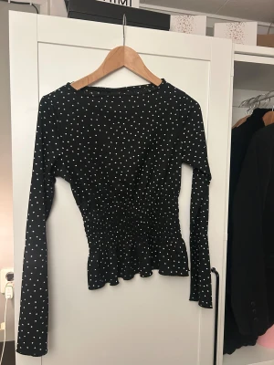 Svart prickig blus från Gina Tricot - Säljer en svart blus från Gina Tricot med vita prickar och smockad midja som ger en snygg siluett. Blusen har långa ärmar och en lätt volang vid nederkanten.