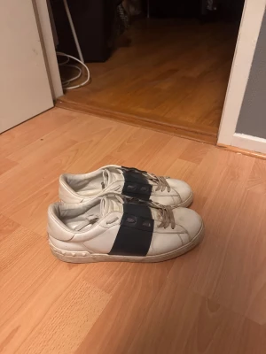 Valentino Garavani vita sneakers - Snygga vita sneakers från Valentino Garavani med bred svart rem över mitten och klassisk snörning. Skorna har rund tå och platt sula, tillverkade i skinn med coola nitar på hälen. Perfekta för dig som gillar exklusiv streetstyle.