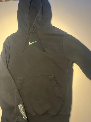Svart Nike hoodie med grön logga - Svart hoodie från Nike med grön Swoosh-logga på både bröstet och ryggen. Tröjan har huva med snörning och en stor magficka framtill. På ärmen finns en extra logga med texten 20/21. Perfekt för dig som gillar sportig och chill stil.