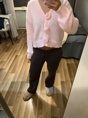 Ljusrosa stickad kofta med blommor - Supergullig ljusrosa stickad kofta med v-ringning och stora dekorativa blommor som knappar framtill. Modellen är croppad med ribbade muddar vid ärmslut och nederkant. Perfekt för dig som gillar söta detaljer och mjuka material.