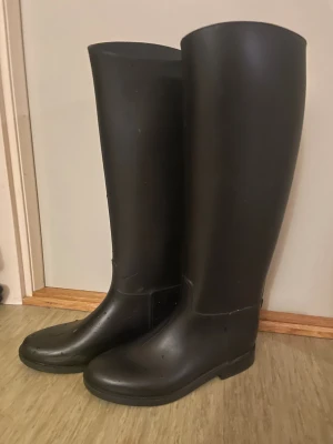 Svarta höga stövlar i syntet - Säljer ett par svarta, höga stövlar med rund tå och platt sula. De är tillverkade i syntetmaterial och har en stilren, minimalistisk look som passar perfekt för regniga dagar eller utomhusaktiviteter. Klassisk design utan extra detaljer.