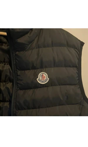 Svart dunväst från Moncler - Svart dunväst från Moncler Grenoble med quiltad design och klassisk logotyp på bröstet. Västen har hög krage, dragkedja framtill och två sidofickor med dragkedja. Perfekt för lager-på-lager och snygg till streetwear eller chill outfits.
