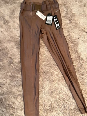 Bruna shimmer tights med bälte från Aim'n - Snygga bruna shimmer tights från Aim'n i storlek S, med ett brett elastiskt bälte och metallmärke vid midjan. Materialet är glansigt och stretchigt, perfekt för träning eller chill. Bältet är avtagbart och har Aim'n-logga. Supersnygg passform och cool detalj.