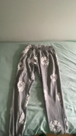 Grå mjukisbyxor Denim Tears M - Grå mjukisbyxor från Denim Tears i storlek M, tillverkade i 100% bomull. Byxorna har ett vitt blommigt mönster och elastisk midja med snörning för skön passform. Perfekta för chill dagar och har en loose fit.