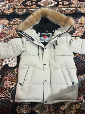 Beige pufferjacka från Zavetti Canada - Snygg beige pufferjacka från Zavetti Canada med stor huva och fuskpäls. Jackan har svarta detaljer, flera fickor med dragkedja och knappar framtill. Insidan är fodrad med svart tyg och logga. Perfekt för kalla dagar och har en riktigt chill vibe.