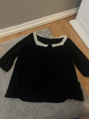 Svart blus med spetskant och knyt - Svart blus med vit spetskant runt halsen och ett sött knyt framtill. Blusen har trekvartsärm och en mjuk, lite lös passform. Perfekt för dig som gillar vintagevibbar och detaljer som sticker ut.