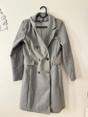 Grå kappa med knappar och krage - Snygg grå kappa med klassisk krage och dubbelknäppning framtill. Jackan har långa ärmar, två fickor med lock och en stilren, rak passform. Perfekt för dig som gillar en clean och tidlös look. Materialet är mjukt och känns varmt, idealiskt för kyliga dagar.