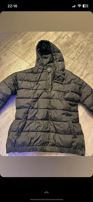 Svart pufferjacka från FB Sister - Svart pufferjacka från FB Sister i storlek M. Jackan har huva med snörning, praktiska fickor och en half zip-dragkedja framtill. Tillverkad i syntetmaterial med quiltad design som håller dig varm under kalla dagar. Perfekt för dig som gillar en clean och sportig look.