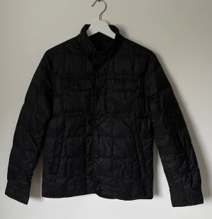 Svart quiltad dunjacka Massimo Dutti - Svart dunjacka från Massimo Dutti med quiltad design och hög krage. Jackan har två bröstfickor med lock, sidofickor och knäppning med knappar. Materialet är polyester med dunfyllning, perfekt för kyliga dagar. Snygg och stilren look som funkar till många outfits. 90% ank dun och 10% fjäder dun. 5H5