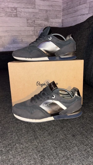 Mörk blåa sneakers från Pepe Jeans - Använts bra men har är lite repade på höger skon men syns inte så värst mycket. Snygga grå sneakers från Pepe Jeans med metallic-silver detaljer på sidorna och vit sula. Skorna har rund tå, snörning och en mix av mocka och syntetmaterial. Perfekt för dig som gillar stilrena och moderna sneakers med coola kontraster.