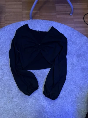 Svart croppad bolerotopp från Monki - Svart croppad bolerotopp från Monki med långärmade puffiga ärmar och snygg rosettdetalj framtill. Toppen har en mjuk, lätt ribbad struktur och är tillverkad i ett stretchigt material som sitter skönt. Perfekt för att lyfta din outfit med en cool touch.