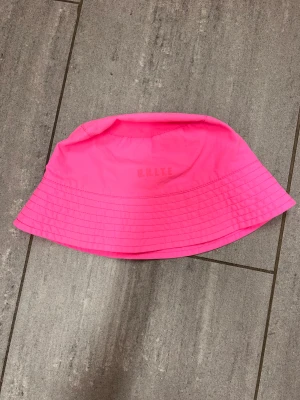 Solhatt  - Bilderna visar en rosa bucket hat från dig UNLTD, inte en väst. Hatten är enfärgad och har en klassisk form med bred monochrom brätte.