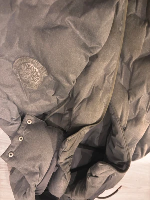 Grå pufferjacka från Messi X SikSilk - Säljer en grå pufferjacka från SikSilk med broderad lejonlogga på bröstet. Jackan har huva med metallöljetter, dragkedja framtill och en snygg quiltad look. Perfekt för kalla dagar när du vill ha både stil och värme.