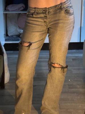 Gråa ripped jeans med raka ben - Säljer ett par gråa jeans med raka ben och snygga slitningar vid knäna. Jeansen har klassisk femficksdesign, knappgylf och låg midja. Materialet är jeans och passformen är normal med lite loose känsla. Perfekt för dig som gillar en avslappnad streetstil.