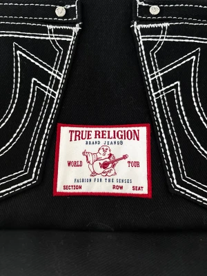 True Religion svart axelväska🖤 - Snygg svart axelväska från True Religion med ikoniska jeansfickor framtill, kontrasterande vit söm och coolt patch med logga. Väskan har både handtag och justerbar axelrem, samt dragkedjor på fickorna. 🖤