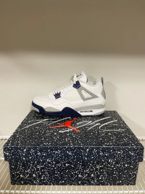 Nike Air Jordan 4 vit och marinblå - Nike Air Jordan 4 sneakers i vit och marinblå med grå detaljer och stänk-mönster på hälen och mellansulan. Skon har snörning, platt sula och meshpaneler på sidorna. Ikonisk Jumpman-logga på baksidan och innersulan. Tillverkad i skinn och syntetmaterial.