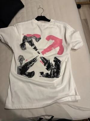 Vit Off-White t-shirt med tryck - Vit t-shirt från Off-White med svart logga och unikt rosa och svart grafiskt tryck både fram och bak. Klassisk passform med rund hals och korta ärmar. Tillverkad i mjuk bomull för en skön känsla. Perfekt för dig som gillar streetwear och vill sticka ut.