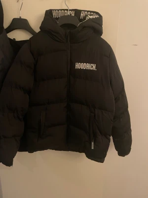 Svart pufferjacka från Hoodrich XL barnstorlek - Svart pufferjacka från Hoodrich i storlek XL med fet logga på bröstet och huvan. Jackan har huva, dragkedja framtill och två sidofickor. Insidan och huvan har Hoodrich-tryck i vitt. Perfekt för dig som gillar streetwear och vill hålla dig varm. Det är storlek XL i barnstorlek passar S perfekt. Pris kan diskuteras. Köpt för 1600kr på kidsbrandstore