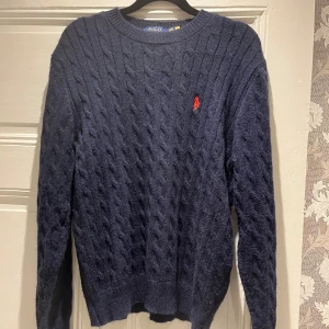 Marinblå stickad tröja från Polo Ralph Lauren - Säljer en marinblå stickad tröja från Polo Ralph Lauren med klassiskt kabelstickat mönster och röd broderad logga på bröstet. Tröjan har rund hals och långa ärmar, perfekt för kyliga dagar.