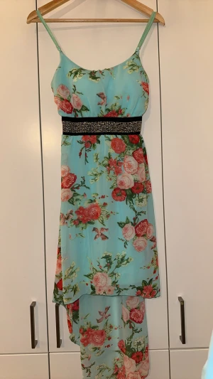Blommig klänning med pärldetaljer - Snygg klänning i ljusblått med blommigt mönster i rosa, rött och grönt. Klänningen har smala axelband och ett svart midjeband dekorerat med pärlor. Materialet är lätt och luftigt, perfekt för varma dagar. Den har en flowy passform och är något längre bak.