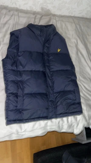 Marinblå dunväst från Lyle & Scott - Snygg marinblå dunväst från Lyle & Scott med klassisk gul logga på bröstet. Västen är quiltad och har hög krage samt dragkedja framtill. Tillverkad i nylon och polyester, perfekt för lager-på-lager under kyliga dagar.
