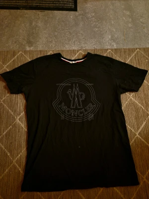 Svart Moncler t-shirt med stor logga - Snygg svart t-shirt från Moncler i storlek XXL men passar som S/M. Stor ton-i-ton Moncler-logga framtill och klassisk patch på ärmen. Rund halsringning med rödvit detalj på insidan av kragen.