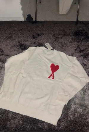 Vit stickad tröja från Ami Paris - Säljer en vit stickad tröja från Ami Paris med ett rött hjärta och bokstaven A broderat på bröstet. Tröjan har rund hals, långa ärmar och ribbade muddar vid ärmslut och nederkant. Perfekt för dig som gillar stilrena detaljer.
