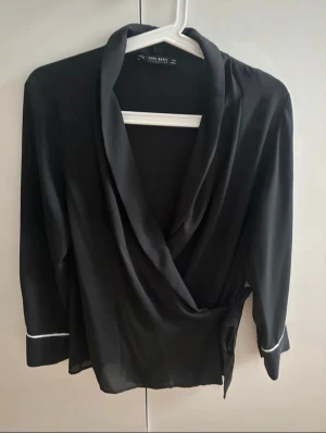 Svart omlott blus/cardigan från Zara Basic - Snygg svart omlottblus från Zara Basic med lång ärm och vit piping vid ärmsluten. Blusen har en snygg draperad krage och knyt i sidan för extra stilpoäng. Perfekt för dig som gillar stilrena och trendiga plagg