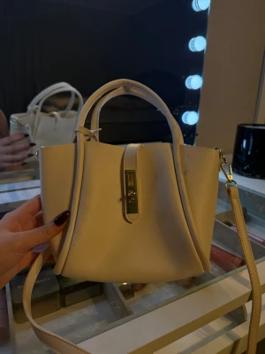 Beige handväska med gulddetalj - Snygg beige handväska i skinnimitation med stilren form och guldfärgad låsdetalj framtill. Väskan har både handtag och avtagbar axelrem, samt ett praktiskt innerfack med dragkedja. Perfekt storlek för det viktigaste och passar till många olika looks.
