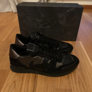 Valentino Rockrunner svarta sneakers - Snygga Valentino Rockrunner sneakers i svart med metallicgrå detaljer och camouflagemönster. Skorna har platt sula med nitar, rund tå och snörning. Materialmix av skinn och textil ger en lyxig känsla. Perfekta för dig som gillar exklusiv streetstyle.