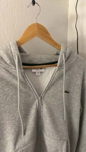 Grå hoodie från Lacoste med dragkedja - Grå hoodie från Lacoste i klassisk passform, med broderad krokodillogga på bröstet. Tröjan har huva med snörning, praktiska fickor och hel dragkedja framtill. Mjuk insida och stilren look, perfekt för chill dagar.
