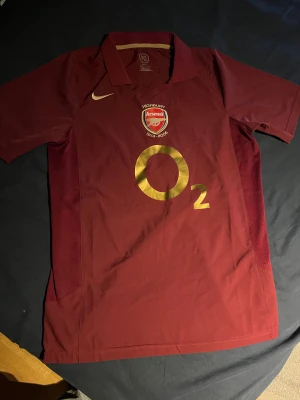 Arsenal Highbury 2005/06 Henry tröja - Säljer en ikonisk Arsenal Highbury 1913-2006 matchtröja med Henry 14 på ryggen. Tröjan är vinröd med guldfärgade detaljer och broderat Arsenal-emblem på bröstet. Tillverkad i polyester, har klassisk krage och korta ärmar. Perfekt för dig som älskar fotboll och Arsenal.