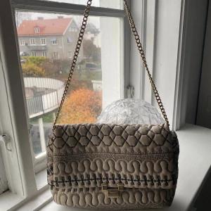 axelväska från Red Valentino - Säljer en beige axelväska från Red valentino med unikt quiltat mönster och snygga svarta broderade detaljer. Väskan har guldfärgad kedja och metallspänne framtill. Det finns lite skador på väskan därav priset som kan skickas i chatten om man är intresserad.