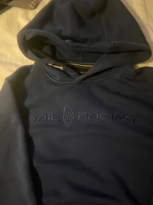 Marinblå Sail Racing hoodie S - Snygg marinblå hoodie från Sail Racing i storlek S. Tröjan har broderad logga på bröstet, huva med snörning och mjukt foder på insidan. Perfekt för dig som gillar sportig och chill stil. Passar dig som vill ha en klassisk hoodie med stilrena detaljer.