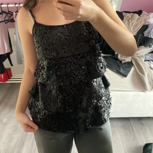 Svart paljettklänning med volanger - Supersnygg svart festtopp med tunna axelband och flera lager volanger. Hela toppen är täckt av glittriga paljetter som ger en festlig look. 
