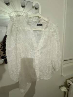 Vit spetsig blus med volang från SHEIN - Supersöt vit blus från SHEIN med transparent spets och fluffiga broderade detaljer. Blusen har volanger vid axlarna, v-ringad hals och knappar framtill. Lätt puffiga långärmar och ett romantiskt uttryck som sticker ut. Perfekt för dig som gillar unika plagg med mycket detaljer.