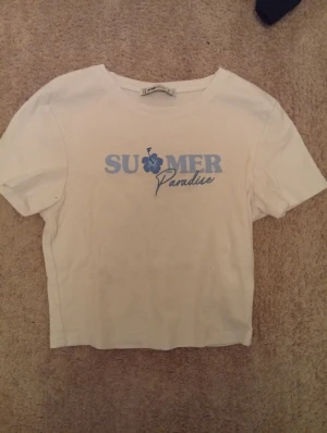 Vit t-shirt med blått tryck FB Sister - Vit t-shirt från FB Sister i storlek M med blått tryck på bröstet där det står 'SUMMER Paradise' och en hibiskusblomma. T-shirten har korta ärmar, rund halsringning och är i mjuk bomull. Perfekt för en chill sommarlook.