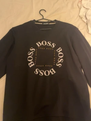 Svart sweatshirt från BOSS - Snygg svart sweatshirt från BOSS med stort vitt BOSS-tryck i cirkel på bröstet och mindre HUGO BOSS-detaljer i guld. Klassisk rund halsringning och långa ärmar. Perfekt för en clean och modern look.