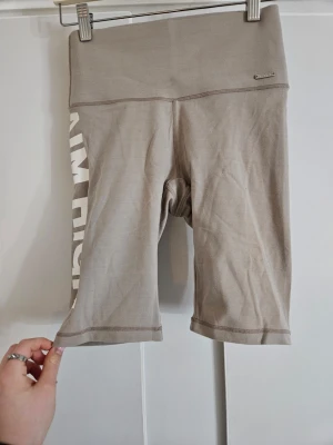 Beige cykelshorts med tryck Aim'n - Snygga beige cykelshorts från Aim'n med hög midja och vitt tryck längs benet där det står 'AIM HIGH.'. Mjukt och stretchigt material som passar perfekt till träning eller sportiga outfits. I storlek S. Knappt använda.