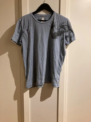 Blå t-shirt med svart tryck från Diesel - Snygg blå t-shirt från Diesel med unikt svart tryck över axeln och bröstet. T-shirten har rund halsringning och korta ärmar. Perfekt för dig som vill ha en cool och avslappnad stil.