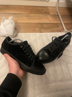 Svarta sneakers från Lanvin i mocka - Säljer ett par svarta Lanvin sneakers med snygga detaljer i mocka och skinn. Skorna har rund tå, platt sula och klassisk snörning. Stilren design med svarta metalldetaljer och svart yttersula. Perfekt för dig som gillar exklusiva och minimalistiska sneakers.
